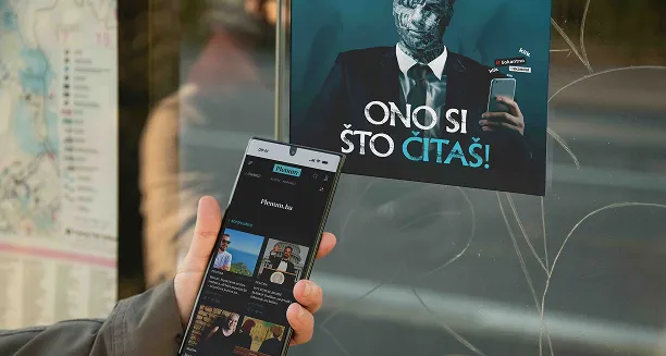 Person holding a smartphone displaying a digital library or reading app, with a poster in the background showing a figure with a textured face and the text 'ONO SI ŠTO ČITAŠ!'