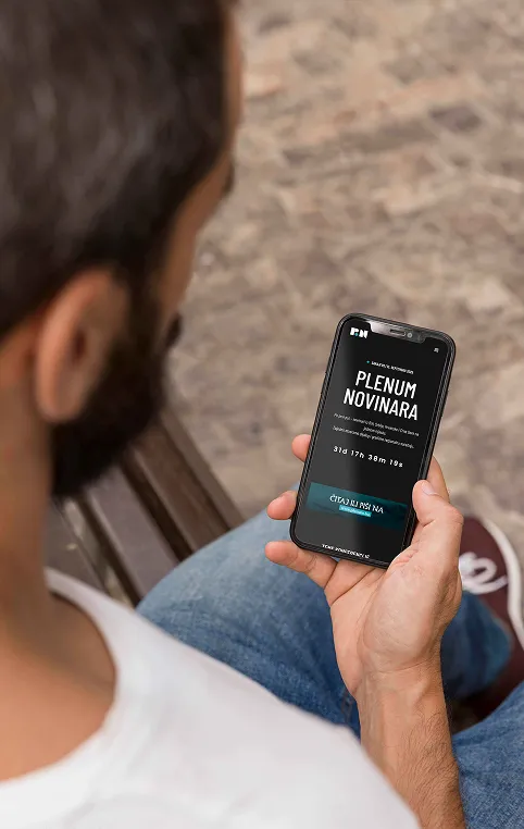 Man sitting outdoors on a bench holding a smartphone displaying a news app in a foreign language.