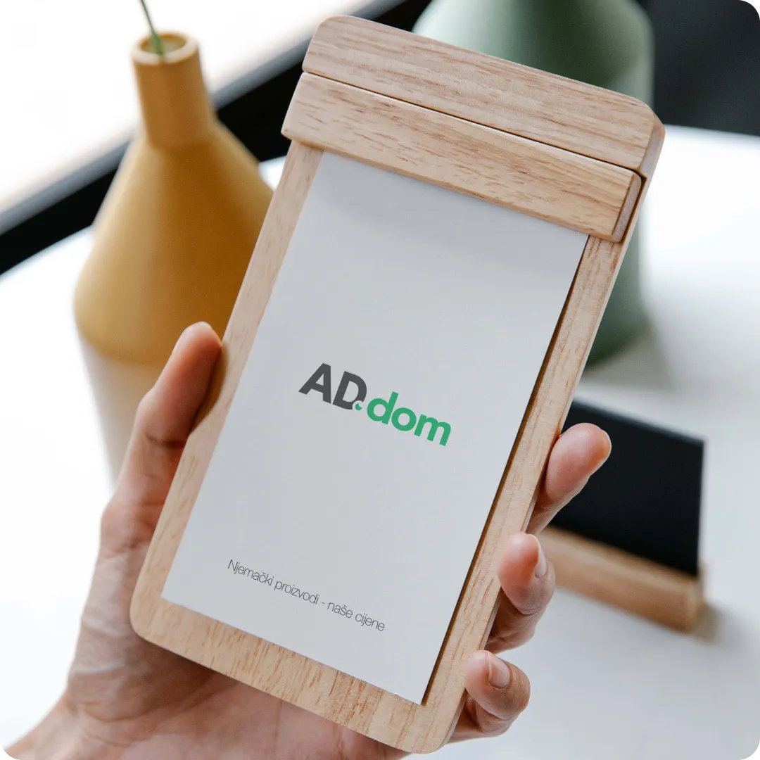 Hand holding a wooden clipboard with a white sheet showing the logo 'AD.dom' and the text 'Njemački proizvodi - naše cijene'.