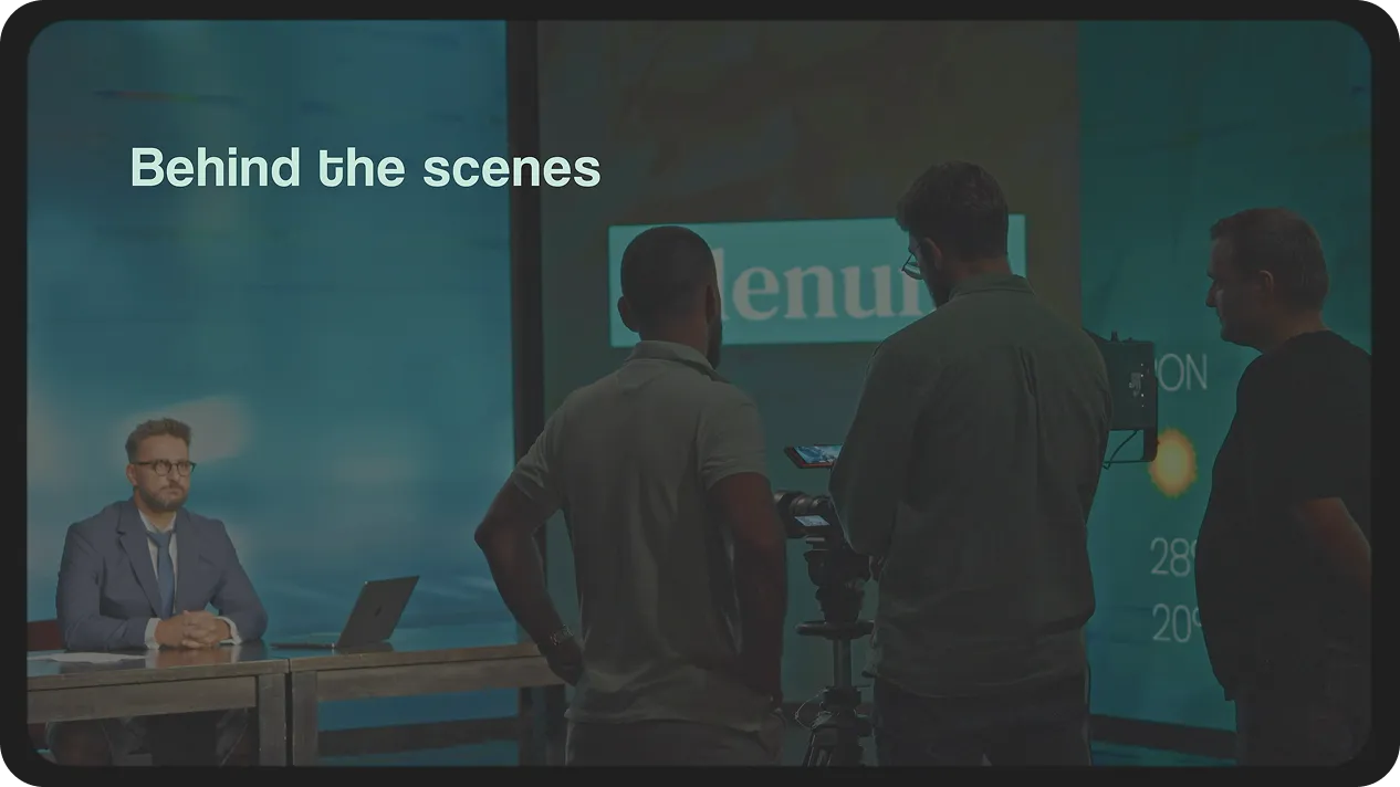 Film crew filming a man in a suit sitting at a desk with a laptop on a set with 'Behind the scenes' text on screen.