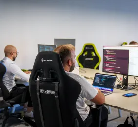 Two men seated at desks working on computers with code displayed on monitors in an office.